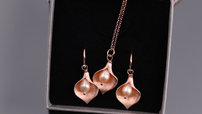 K&ouml;nnte beinhalten: Ein Schmuckset bestehend aus einer Halskette und zwei Ohrh&auml;ngern in Ros&eacute;gold-Optik. Jedes St&uuml;ck zeigt ein strukturiertes, mattes Calla-Lilien-Bl&uuml;tendesign mit einer einzelnen, glatten, cremefarbenen Perle, die in der Mitte des Bl&uuml;tenblatts sitzt.