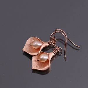 K&ouml;nnte beinhalten: Ein Paar ohrringe in ros&eacute;goldfarbener Optik in Form von Calla-Lilien, jeweils mit einer zentralen wei&szlig;en Perle. Die Metallbl&auml;tter haben eine geb&uuml;rstete, matte Textur und die Ohrringe sind an geschwungenen Ohrhaken befestigt.