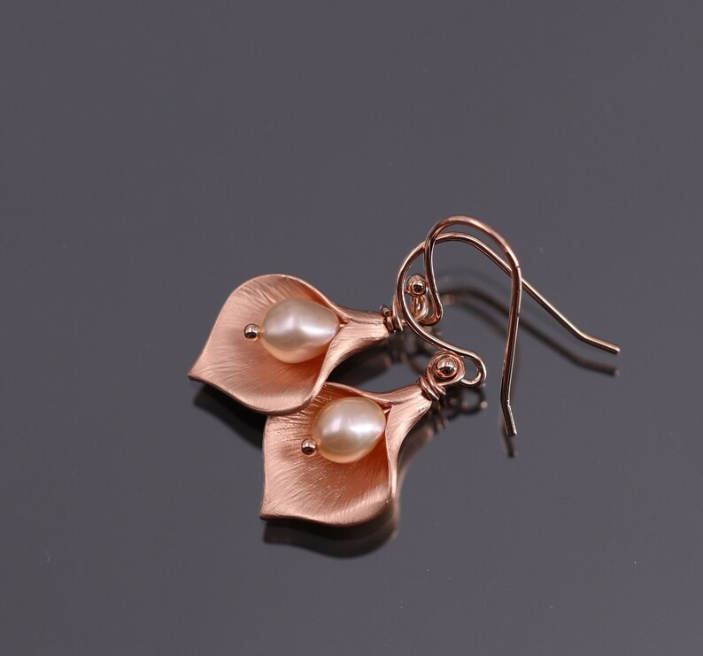K&ouml;nnte beinhalten: Ein Paar ohrringe in ros&eacute;goldfarbener Optik in Form von Calla-Lilien, jeweils mit einer zentralen wei&szlig;en Perle. Die Metallbl&auml;tter haben eine geb&uuml;rstete, matte Textur und die Ohrringe sind an geschwungenen Ohrhaken befestigt.