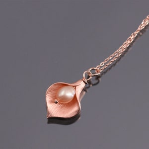 K&ouml;nnte beinhalten: Eine ros&eacute;goldfarbene Halskette mit einem Anh&auml;nger in Form einer Calla-Lilienbl&uuml;te mit einer texturierten, geb&uuml;rsteten Metalloberfl&auml;che. Eine einzelne, glatte, oval geformte cremefarbene Perle ist in der Mitte der Bl&uuml;te eingesetzt. Der Anh&auml;nger h&auml;ngt an einer zarten Kette.