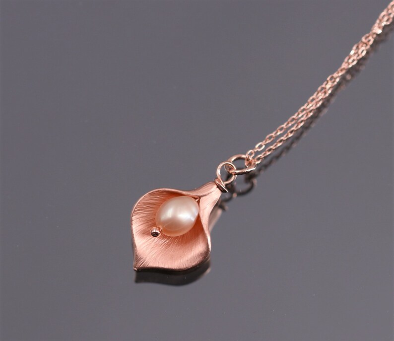 K&ouml;nnte beinhalten: Eine ros&eacute;goldfarbene Halskette mit einem Anh&auml;nger in Form einer Calla-Lilienbl&uuml;te mit einer texturierten, geb&uuml;rsteten Metalloberfl&auml;che. Eine einzelne, glatte, oval geformte cremefarbene Perle ist in der Mitte der Bl&uuml;te eingesetzt. Der Anh&auml;nger h&auml;ngt an einer zarten Kette.