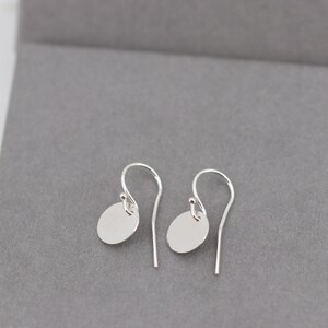 Op de afbeelding: Een paar zilveren oorhangers met een minimalistisch ontwerp. Elke oorbel heeft een kleine, platte, ovale bedel die aan een gebogen oorhaak hangt. De oorbellen worden gepresenteerd op een grijze ondergrond.