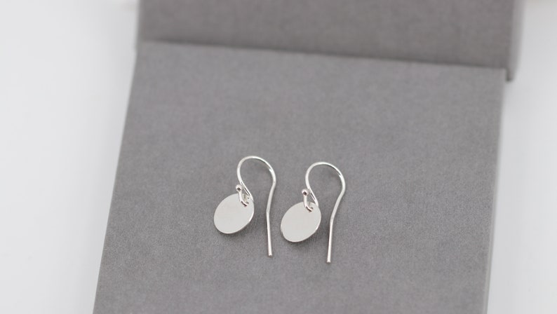 Op de afbeelding: Een paar zilveren oorhangers met een minimalistisch ontwerp. Elke oorbel heeft een kleine, platte, ovale bedel die aan een gebogen oorhaak hangt. De oorbellen worden gepresenteerd op een grijze ondergrond.