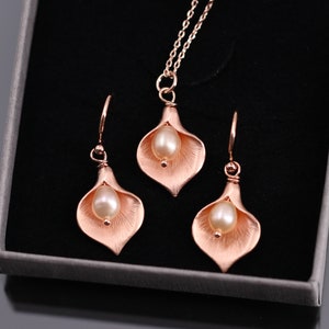 K&ouml;nnte beinhalten: Ein Schmuckset bestehend aus einer Halskette und zwei Ohrringen, jeweils mit einem ros&eacute;goldfarbenen Calla-Lilien-Anh&auml;nger und einer cremefarbenen Perle in der Mitte. Die Halskette enth&auml;lt eine passende Kette, alles pr&auml;sentiert in einer Schachtel.