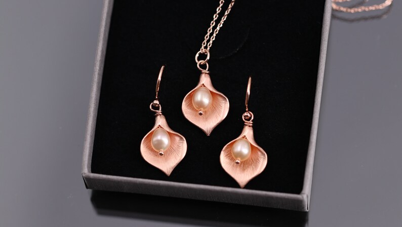K&ouml;nnte beinhalten: Ein Schmuckset bestehend aus einer Halskette und zwei Ohrringen, jeweils mit einem ros&eacute;goldfarbenen Calla-Lilien-Anh&auml;nger und einer cremefarbenen Perle in der Mitte. Die Halskette enth&auml;lt eine passende Kette, alles pr&auml;sentiert in einer Schachtel.