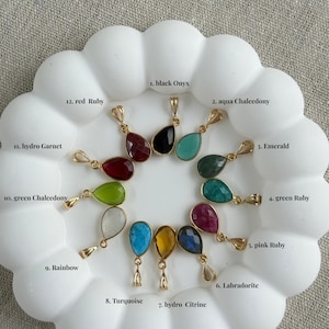 Può includere: Dodici ciondoli in pietra preziosa a forma di goccia con ganci dorati, disposti in cerchio. Etichettati 1. black Onyx, 2. aqua Chalcedony, 3. Emerald, 4. green Ruby, 5. pink Ruby, 6. Labradorite, 7. hydro Citrine, 8. Turquoise, 9. Rainbow, 10. green Chalcedony, 11. hydro Garnet, 12. red Ruby.