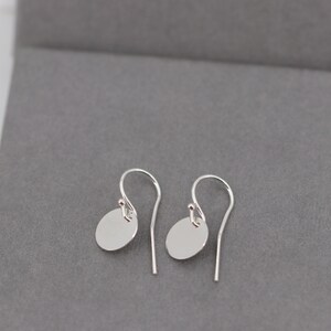 Op de afbeelding: Een paar zilveren oorhangers. Elke oorbel heeft een kleine, platte, cirkelvormige hanger die aan een gebogen oorhaak hangt. De oorbellen worden gepresenteerd op een grijze ondergrond.