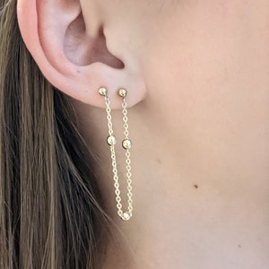 Könnte beinhalten: Goldene Kettenohrringe mit kleinen runden Perlen. Die Kette ist an einem kleinen Ohrstecker befestigt.