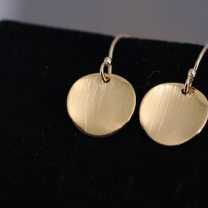 Könnte beinhalten: Ein Paar goldfarbene Ohrhänger mit gebürsteter Oberfläche. Die Ohrringe sind rund und haben ein schlichtes Design.