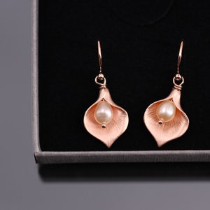 K&ouml;nnte beinhalten: Ein Paar Ohrh&auml;nger in Ros&eacute;gold-Optik in Form von Calla-Lilien. Jeder Ohrring verf&uuml;gt &uuml;ber eine mittig platzierte, cremefarbene Perle, die in die texturierte, bl&uuml;tenblattartige Metallstruktur der Blume eingebettet ist.