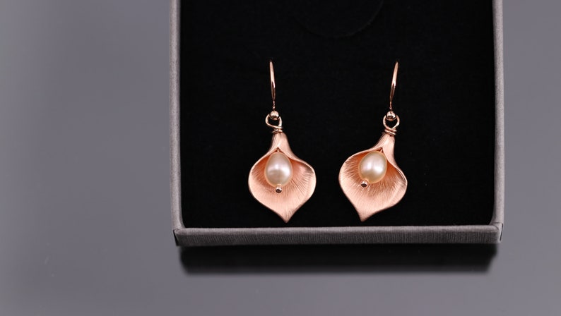 K&ouml;nnte beinhalten: Ein Paar Ohrh&auml;nger in Ros&eacute;gold-Optik in Form von Calla-Lilien. Jeder Ohrring verf&uuml;gt &uuml;ber eine mittig platzierte, cremefarbene Perle, die in die texturierte, bl&uuml;tenblattartige Metallstruktur der Blume eingebettet ist.