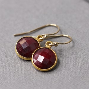 Puede incluir: Un par de pendientes colgantes dorados con piedras preciosas redondas, facetadas, de color rojo oscuro. Los pendientes tienen un diseño sencillo con cierre de gancho. Las piedras preciosas están engastadas en un bisel dorado.