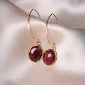 Puede incluir: Un par de pendientes de aro dorados con un colgante de piedra preciosa roja oscura. Los pendientes están sobre un fondo de tela rosa claro.