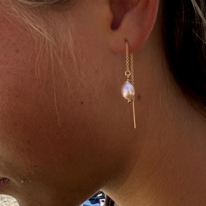 May include: A close-up of a gold-colored earring with a chain and a dangling, light pink pearl. The earring also features a small, gold-colored bar. The earring is worn by a person with blonde hair.
