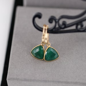 Peut inclure: Une paire de boucles d'oreilles créoles en or avec des gouttes triangulaires en pierre précieuse verte. Les boucles d'oreilles sont présentées sur une surface grise.