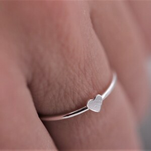 Könnte beinhalten: Ein silberner Ring mit einem herzförmigen Design. Der Ring wird an einem Finger getragen.