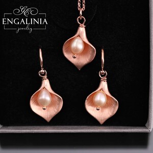 K&ouml;nnte beinhalten: Ein Schmuckset bestehend aus einer Halskette und zwei Ohrringen in Ros&eacute;gold-Optik, jeweils in Form einer Calla-Lilie mit einer zentralen wei&szlig;en Perle. Die St&uuml;cke liegen in einer Schachtel mit dem Schriftzug ENGALINIA jewelry oben links.