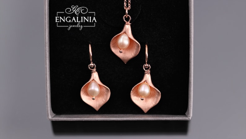K&ouml;nnte beinhalten: Ein Schmuckset bestehend aus einer Halskette und zwei Ohrringen in Ros&eacute;gold-Optik, jeweils in Form einer Calla-Lilie mit einer zentralen wei&szlig;en Perle. Die St&uuml;cke liegen in einer Schachtel mit dem Schriftzug ENGALINIA jewelry oben links.