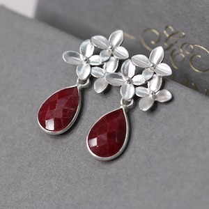 Könnte beinhalten: Silberne Ohrringe mit einem Cluster aus kleinen weißen Blütenanhängern und einem roten, tropfenförmigen Edelstein.