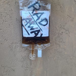 May include: A transparent plastic blood bag containing a dark brown liquid. The bag is marked with the word "RAD" in black marker. A label with printed text is visible, along with a clear attached tube.