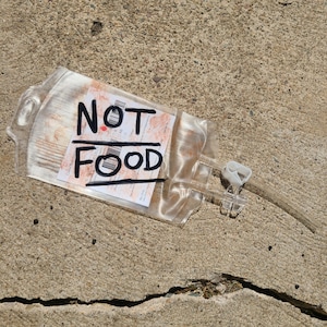 May include: A clear plastic medical IV bag with a white label featuring red blood-like splatters and the handwritten black text NOT FOOD. The bag includes two clear plastic tubes and a white plastic clamp attached to the side of the container.
