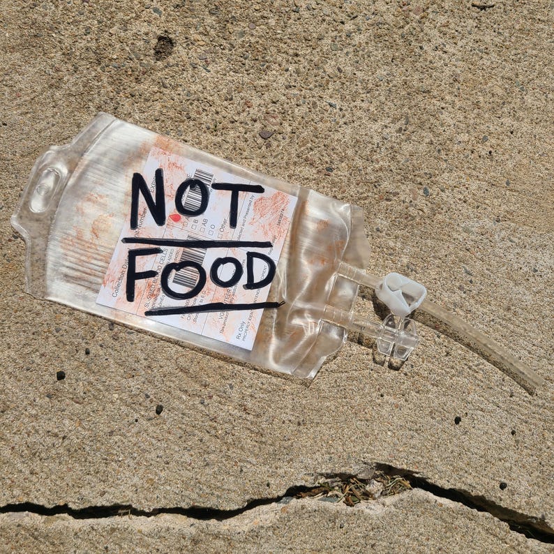 May include: A clear plastic medical IV bag with a white label featuring red blood-like splatters and the handwritten black text NOT FOOD. The bag includes two clear plastic tubes and a white plastic clamp attached to the side of the container.
