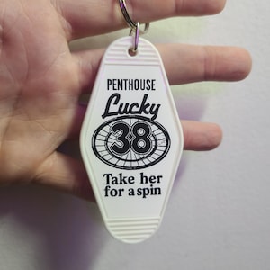 Puede incluir: Llavero blanco estilo motel con el texto "Penthouse Lucky 38" y "Take her for a spin" en negro. El llavero tiene un anillo de metal plateado en la parte superior.