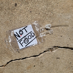 May include: A clear, empty plastic medical blood bag featuring a white label with black text and barcodes. The words NOT FOOD are written in large, bold black marker across the front. The bag includes a clear plastic tube and a white clamp.