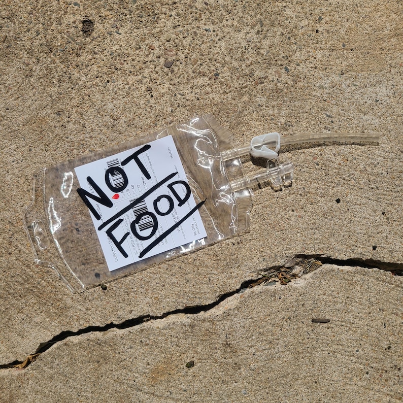 May include: A clear, empty plastic medical blood bag featuring a white label with black text and barcodes. The words NOT FOOD are written in large, bold black marker across the front. The bag includes a clear plastic tube and a white clamp.