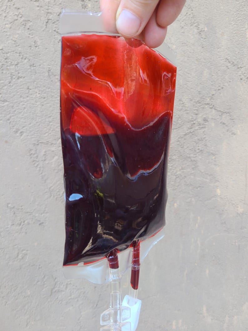 May include: A clear plastic bag filled with a dark red liquid, possibly blood. The liquid is layered, with a brighter red at the top and a darker shade at the bottom. Two tubes extend from the bottom of the bag. The bag is held by a person.