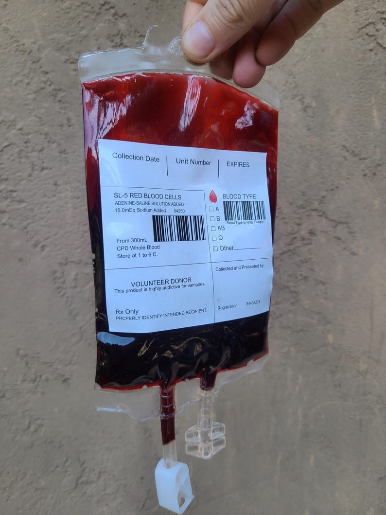 May include: A clear plastic bag filled with a dark red liquid, labelled "SL-5 RED BLOOD CELLS." The bag includes a barcode, blood type options, and text indicating it is from a volunteer donor. The bag is held by a hand.