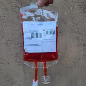 May include: A clear plastic bag filled with a red liquid, resembling a blood bag. The bag has a label with text including "SL-5 RED BLOOD CELLS" and "BLOOD TYPE". Two tubes extend from the bottom, with a white and clear plastic connector.