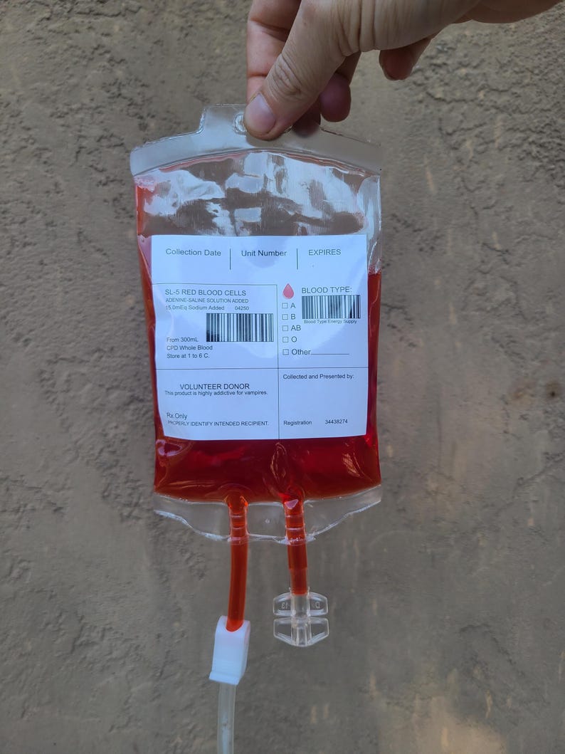 May include: A clear plastic bag filled with a red liquid, resembling a blood bag. The bag has a label with text including "SL-5 RED BLOOD CELLS" and "BLOOD TYPE". Two tubes extend from the bottom, with a white and clear plastic connector.