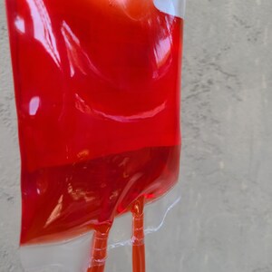 May include: A transparent plastic bag filled with a vibrant red liquid, similar to an IV drip bag. Two clear tubes emerge from the base, secured by a transparent plastic clip. The bag is set against a textured, light grey backdrop.