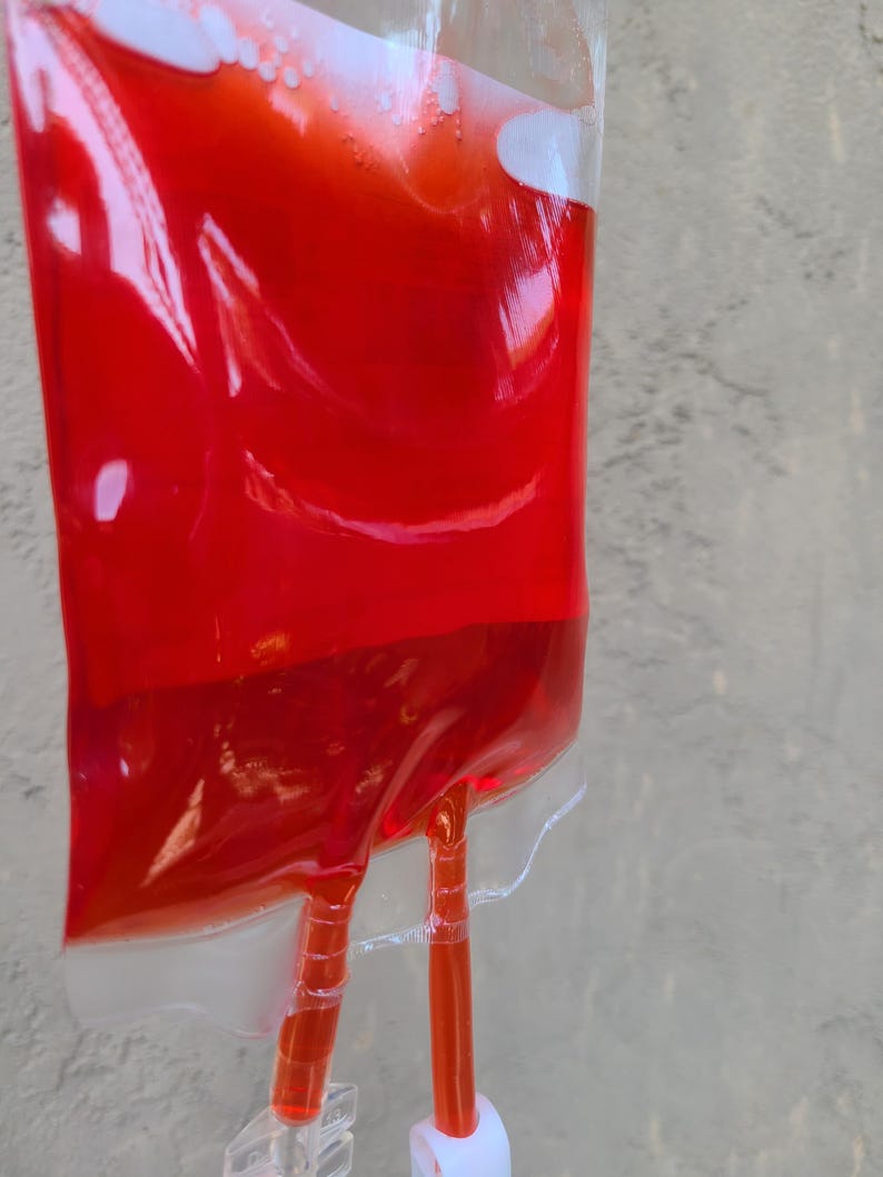 May include: A transparent plastic bag filled with a vibrant red liquid, similar to an IV drip bag. Two clear tubes emerge from the base, secured by a transparent plastic clip. The bag is set against a textured, light grey backdrop.