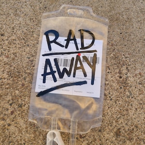 May include: A clear, empty plastic medical blood bag featuring a white label with black text and a barcode. The words RAD AWAY are written in large, bold black marker across the center of the bag, partially obscuring the printed label details.