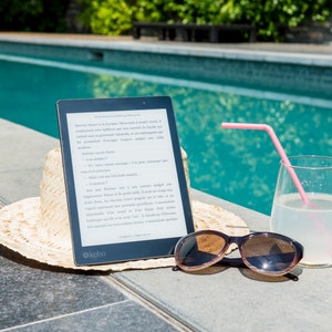 A black e-reader with a white screen displaying an ebook, a straw hat, sunglasses, and a glass of lemonade with a pink straw on a gray tiled surface by a swimming pool. These books represent the ebooks that are included in this downloadable pack of rare witchcraft, magical, spell craft, and paganism books.