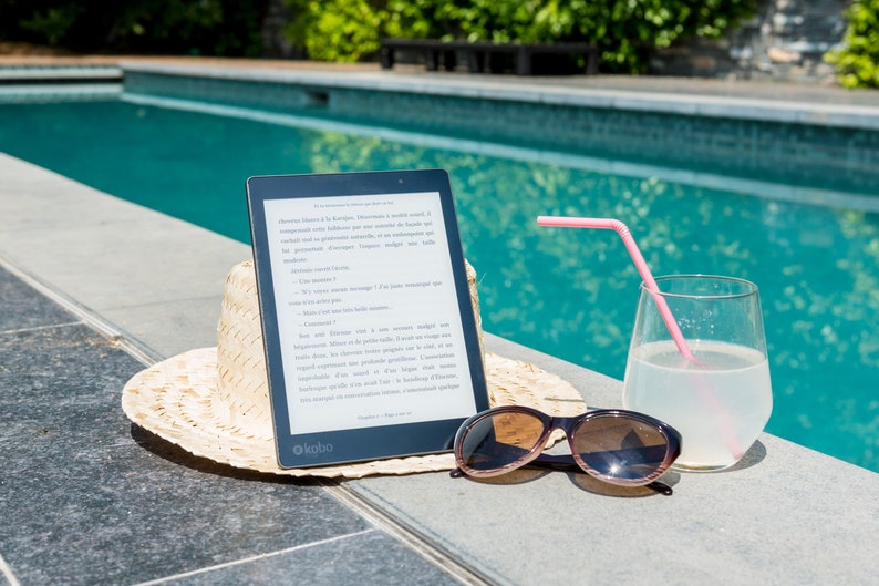 A black e-reader with a white screen displaying an ebook, a straw hat, sunglasses, and a glass of lemonade with a pink straw on a gray tiled surface by a swimming pool. These books represent the ebooks that are included in this downloadable pack of rare witchcraft, magical, spell craft, and paganism books.