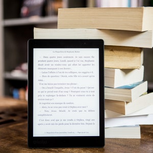 A black and white e-reader with a white screen displaying a novel. The e-reader is sitting on a wooden table next to a stack of books. These books represent the ebooks that are included in this downloadable pack of rare witchcraft, magical, spell craft, and paganism books.