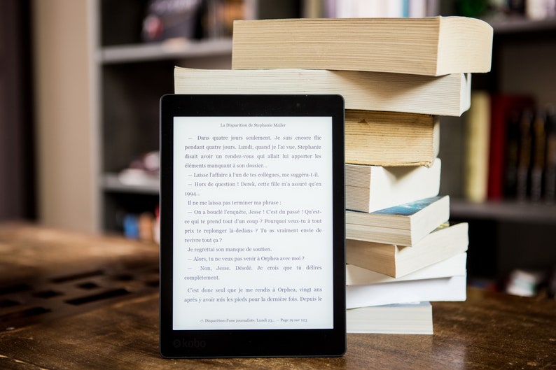 A black and white e-reader with a white screen displaying a novel. The e-reader is sitting on a wooden table next to a stack of books. These books represent the ebooks that are included in this downloadable pack of rare witchcraft, magical, spell craft, and paganism books.