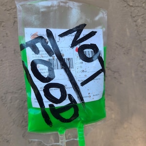 May include: A clear plastic IV bag filled with bright green liquid. The bag has a label with illegible text and is marked with large black marker strokes. The bag is held up by a hand.