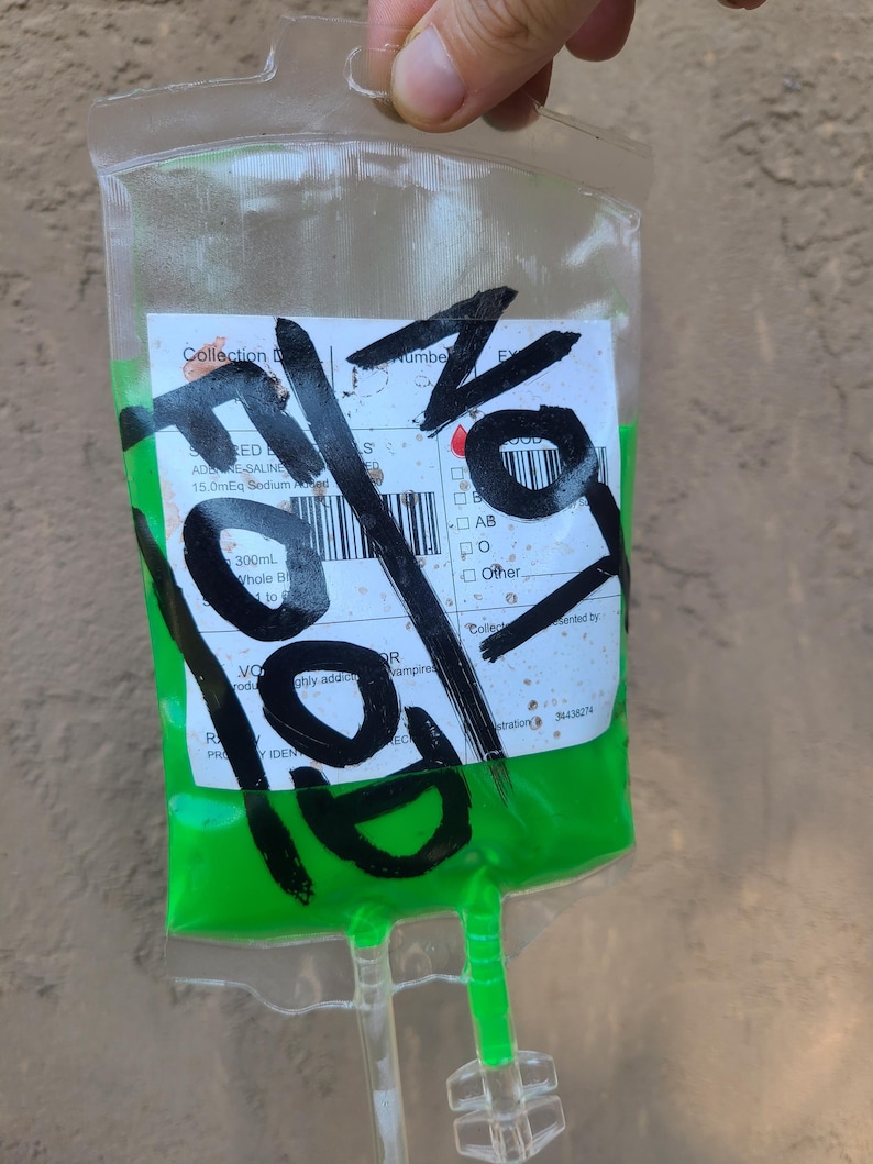 May include: A clear plastic IV bag filled with bright green liquid. The bag has a label with illegible text and is marked with large black marker strokes. The bag is held up by a hand.