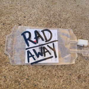 May include: A clear, flexible plastic medical blood bag featuring a white rectangular label with black text and barcodes. The words RAD AWAY are written in large, bold black marker across the center of the bag, partially obscuring the label.