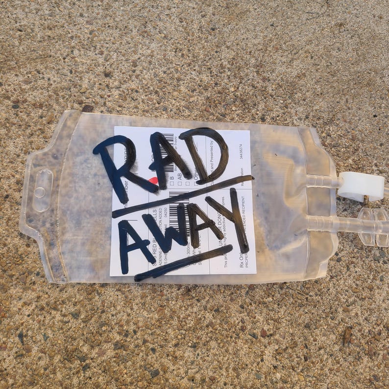 May include: A clear, flexible plastic medical blood bag featuring a white rectangular label with black text and barcodes. The words RAD AWAY are written in large, bold black marker across the center of the bag, partially obscuring the label.
