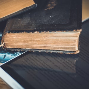 A close-up of three books stacked on top of each other.  These books represent the ebooks that are included in this downloadable pack of rare witchcraft, magical, spell craft, and paganism books. The top book is a thick, black leather-bound book with gold lettering on the spine. The middle book is a brown leather-bound book with a gold spine. The bottom book is a paperback book with a blue cover. A white tablet computer is lying on top of the books.