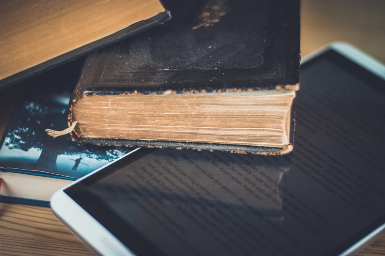A close-up of three books stacked on top of each other.  These books represent the ebooks that are included in this downloadable pack of rare witchcraft, magical, spell craft, and paganism books. The top book is a thick, black leather-bound book with gold lettering on the spine. The middle book is a brown leather-bound book with a gold spine. The bottom book is a paperback book with a blue cover. A white tablet computer is lying on top of the books.