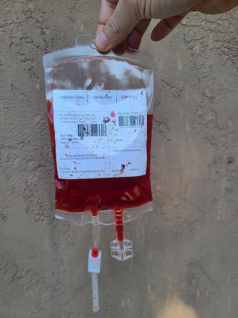 May include: A clear plastic blood bag filled with red liquid, held by a hand. The bag has attached tubes and printed labels with text including "SL-5 RED BLOOD CELLS" and "BLOOD TYPE".