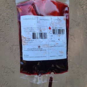 May include: A transparent plastic bag filled with a dark red liquid, resembling blood. The bag has a label with text including "SL-5 RED BLOOD CELLS" and "VOLUNTEER DONOR". The bag has a clear tube and a white label with text.