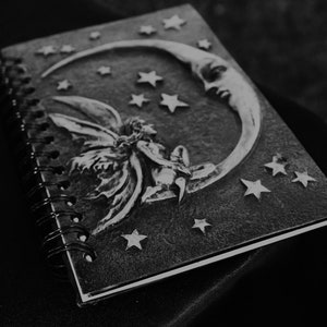 A black and white journal with a spiral binding. The cover features a silver embossed design of a fairy sitting on a crescent moon with stars. This book represents the ebooks that are included in this downloadable pack of rare witchcraft, magical, spell craft, and paganism books.