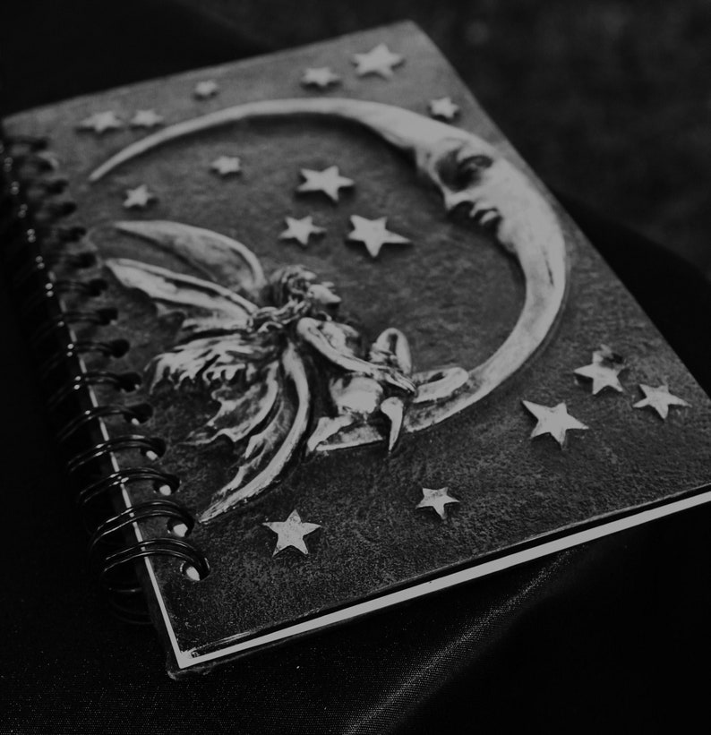 A black and white journal with a spiral binding. The cover features a silver embossed design of a fairy sitting on a crescent moon with stars. This book represents the ebooks that are included in this downloadable pack of rare witchcraft, magical, spell craft, and paganism books.